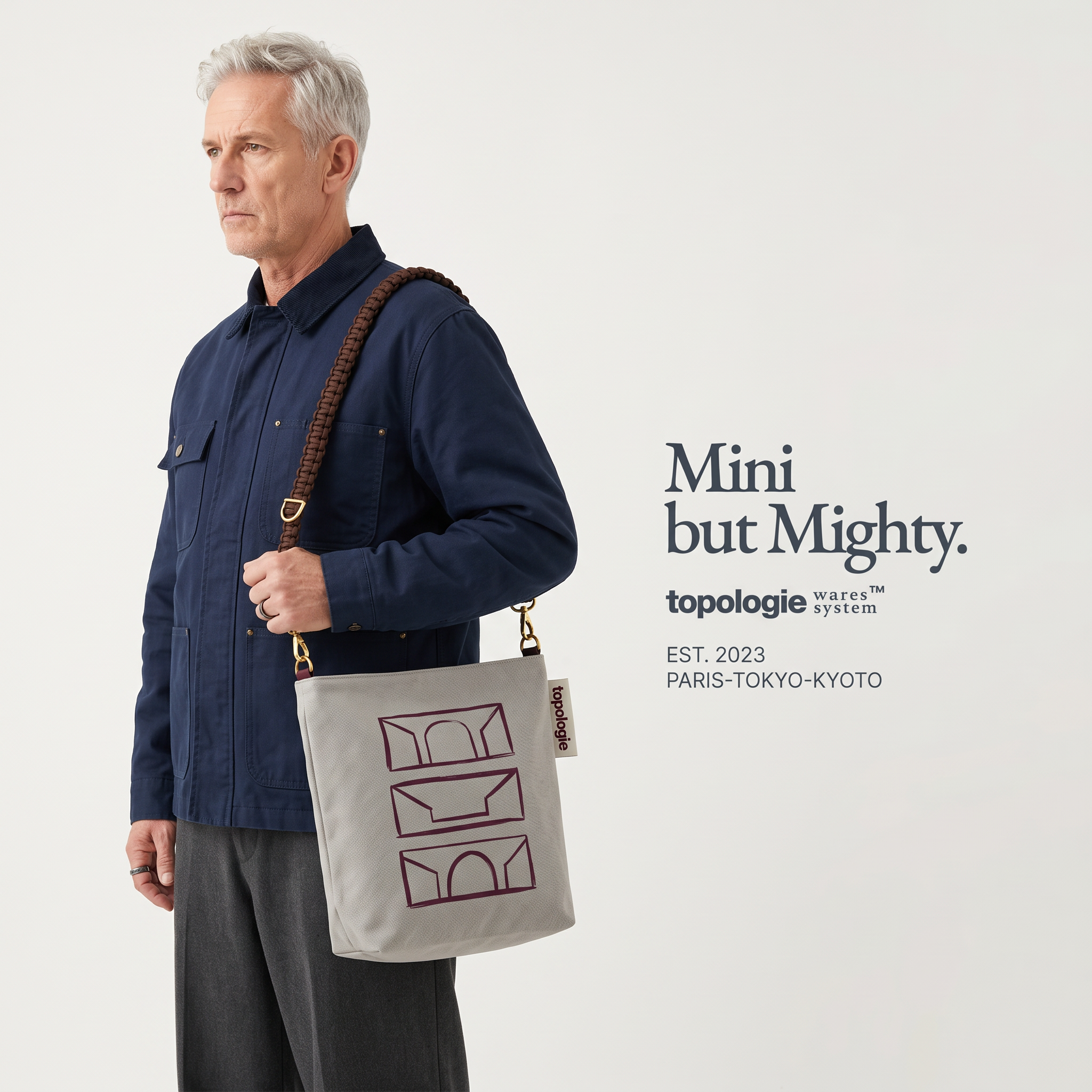 Silver-haired woman in profile in a charcoal jacket, carrying a canvas Topologie tote with a padded red strap