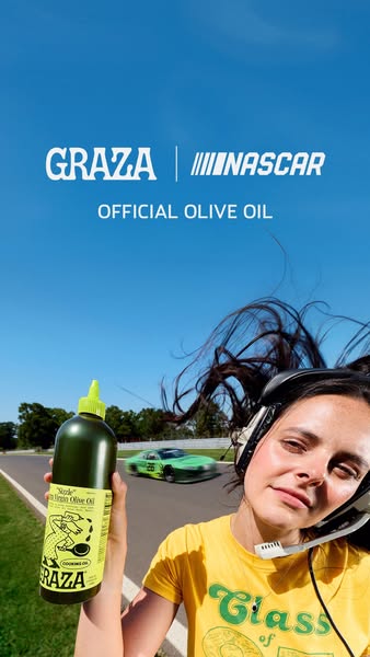 Woman in headset with hair blowing holds Graza Sizzle olive oil as a green race car passes on a track under blue sky