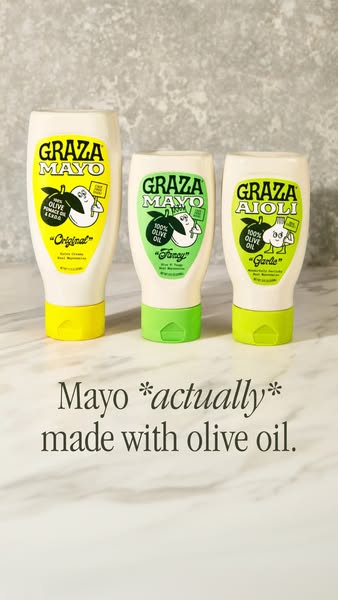 Three Graza squeeze bottles, Mayo Original, Mayo Fancy and Garlic Aioli, on white marble against a grey stone wall