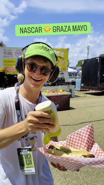 Smiling fan in Graza cap and NASCAR lanyard squeezes Graza Mayo onto a hot dog at a racetrack food stand