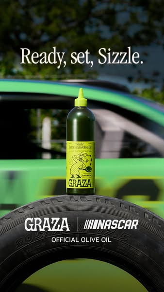 Graza Sizzle olive oil bottle standing on a black race tyre with a green car speeding behind, text Ready, set, Sizzle