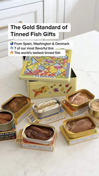 Seven open Fishwife tins of fish arranged around a yellow stained-glass gift tin on a white marble kitchen counter