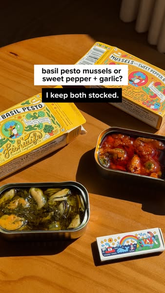 Two open tins of mussels, basil pesto and sweet pepper with garlic, with their yellow boxes on a sunny wooden table