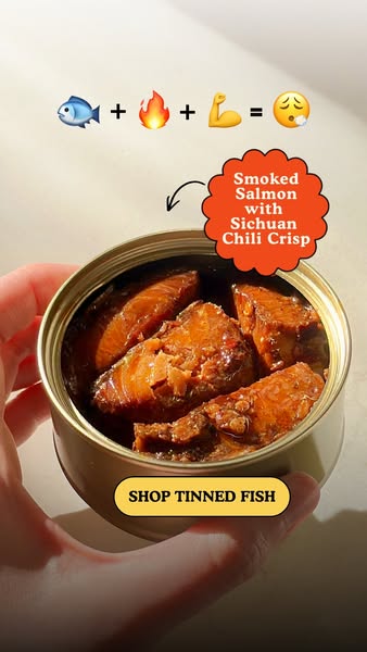 Hand holding an open tin of smoked salmon in chili crisp with an emoji equation and orange label badge above