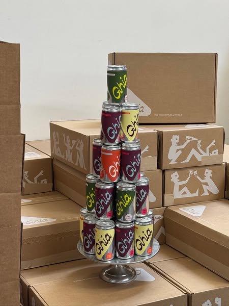 Pyramid of Ghia Le Spritz cans on a silver cake stand surrounded by kraft shipping boxes with toasting illustrations