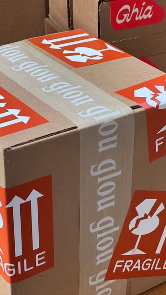 Close-up of cardboard box with white glou glou printed tape, orange fragile and this-side-up stickers and red Ghia sticker