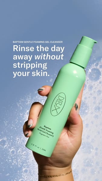 Hand with painted nails holding a mint-green Dieux Baptism cleanser bottle against blue with foam bubbles