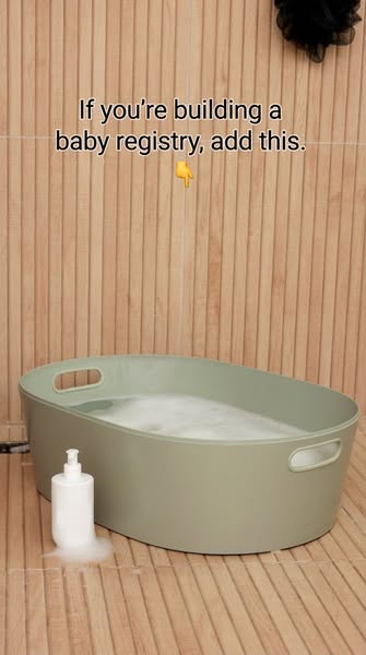 Sage green Lalo baby bath tub filled with bubbles beside a soap bottle, against a slatted wood wall and floor