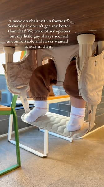 Under-table view of a toddler in white socks resting feet on the footrest of a cream Lalo hook-on chair at a restaurant