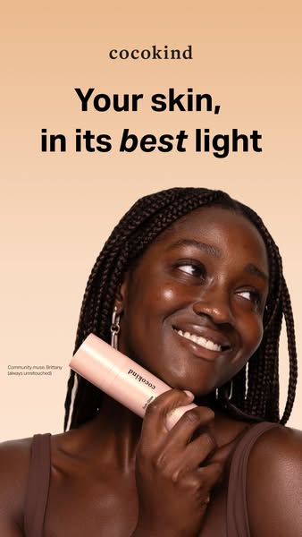 Smiling woman with braids holding a Cocokind Soft-Glow Illuminator to her cheek on a peach background