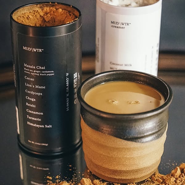 Open black MUD WTR tin with ingredient list next to a handmade ceramic cup of creamy chai and white creamer tin