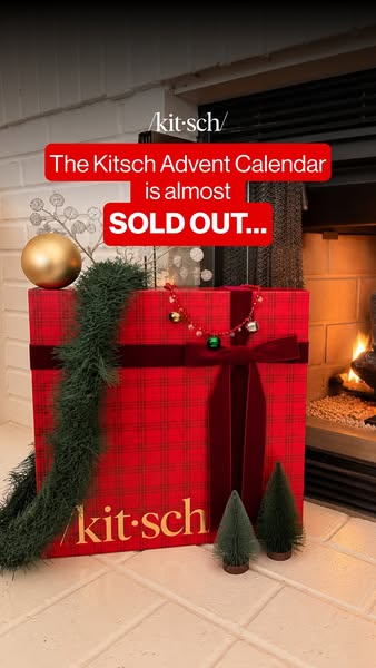 Large red plaid Kitsch advent calendar box with velvet bow beside a lit fireplace, text says it is almost sold out