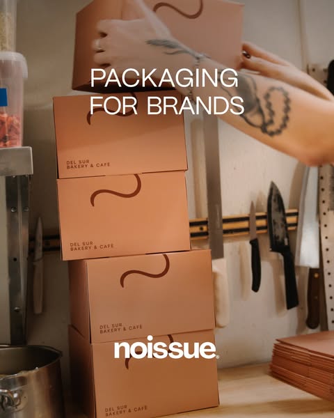 Noissue ad: a tattooed arm stacking five terracotta Del Sur Bakery & Café boxes in a busy café kitchen, a magnetic knife…