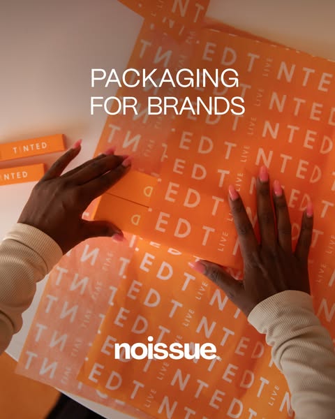 Noissue ad: a top-down shot of manicured hands wrapping a small Tinted product box in orange branded tissue paper