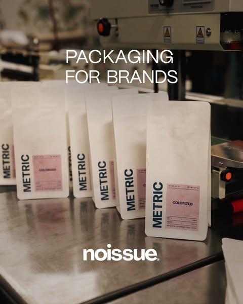 Noissue ad: a row of white Metric coffee bags lined up on a stainless steel table next to a sealing machine in a roastery