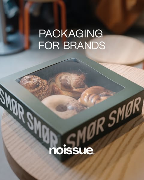 Noissue ad: a forest-green SMØR pastry box with a clear lid showing cinnamon buns and a bagel, set on a light wooden café…