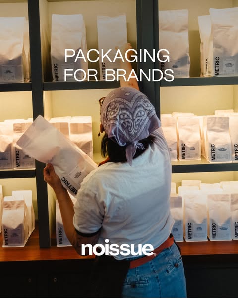 Noissue ad: a barista in a bandana stocking a backlit wooden shelf with rows of white Metric coffee bags