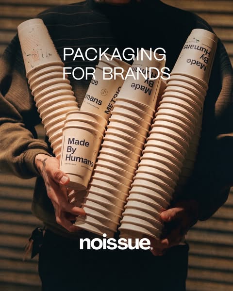 Noissue ad: a man in a knit sweater holding four tall stacks of cream Made By Humans takeaway cups against a dark slatted…