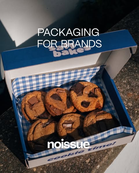 Noissue ad: an open blue-and-white cookie box lined with blue gingham paper, holding six chunky chocolate chip cookies in…