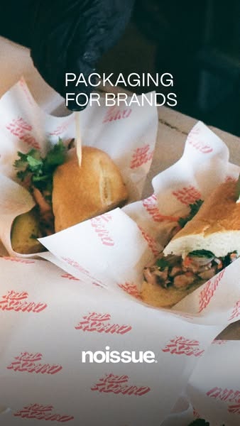 Noissue ad: a gloved hand holding a cut baguette sandwich wrapped in white paper printed with a red script logo
