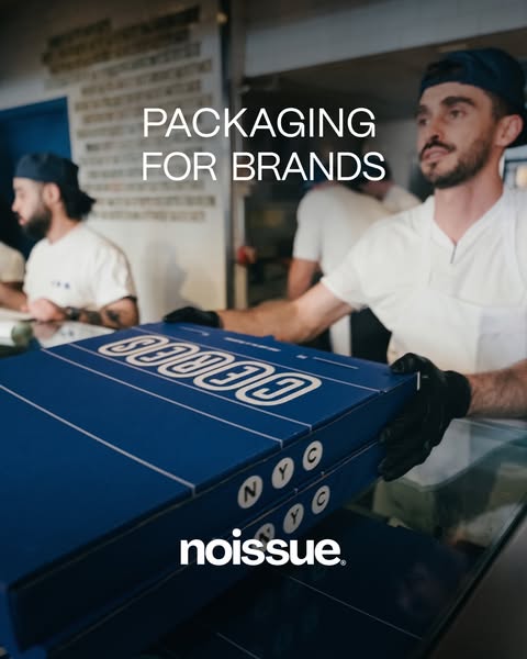 Noissue ad: pizza makers in white tees and caps sliding stacked blue Ceres NYC pizza boxes across a counter
