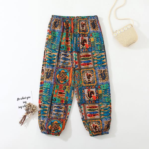 Flat lay of colorful boho patchwork harem pants with elastic cuffs, straw bag and dried flowers on white background