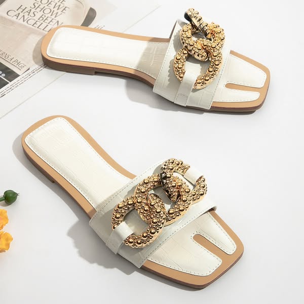 White croc-effect flat slide sandals with chunky gold chain detail on white surface with magazine and yellow flowers