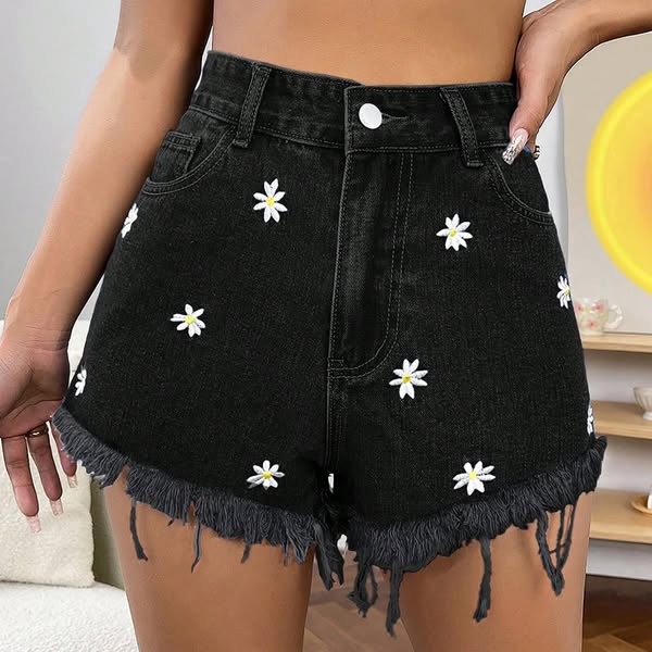 Close-up of model wearing black high-waisted denim shorts with white daisy embroidery and frayed hem in a cozy room