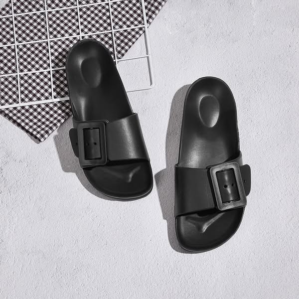 Top-down view of black buckle slide sandals with contoured footbed on grey concrete with gingham cloth and wire grid