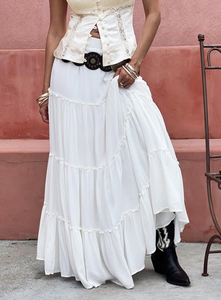Model in a white tiered maxi skirt, cream corset top, black concho belt and cowboy boots against a terracotta wall