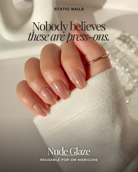 Hand in a white fleece sleeve wearing glossy sheer nude press-on nails with a thin gold ring in a bright minimal room