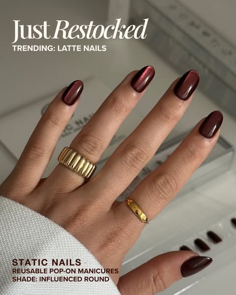 Hand with gold rings wearing glossy deep burgundy chrome press-on nails, with Static Nails packaging blurred behind