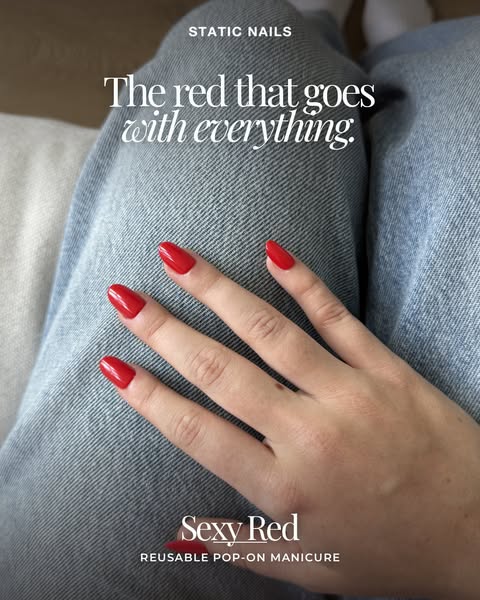 Hand with glossy classic red almond press-on nails resting on light blue denim jeans, with a pale sofa in the background