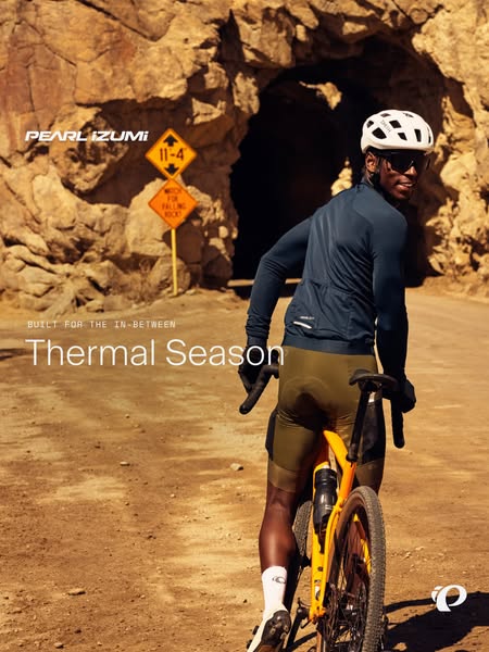Gravel cyclist in navy thermal jersey and olive bibs on orange bike looking back by rock tunnel, headline Thermal Season