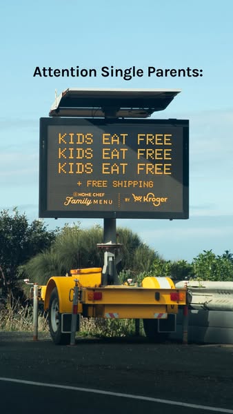 Yellow trailer-mounted LED road sign under a blue sky repeating Kids Eat Free three times for single parents