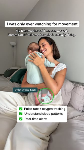 Smiling mom on a bed cuddles her baby, whose foot wears a circled mint Owlet Dream Sock, with three checkmarked benefits