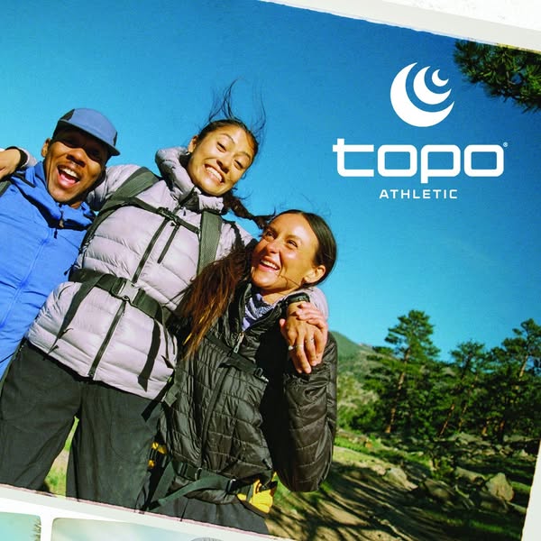 Three laughing hikers in puffer jackets and backpacks hugging under a blue sky with the white Topo Athletic logo