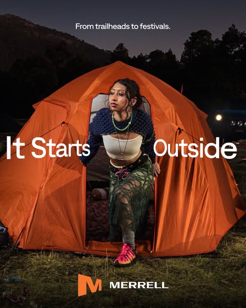 Woman in knit cardigan, crop top and lace skirt steps out of a lit orange dome tent at night wearing pink Merrell shoes