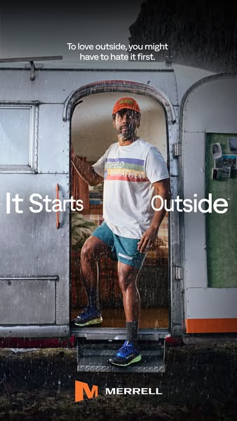 Man in orange cap, striped tee and shorts steps out of a silver trailer into the rain in blue Merrell trail shoes