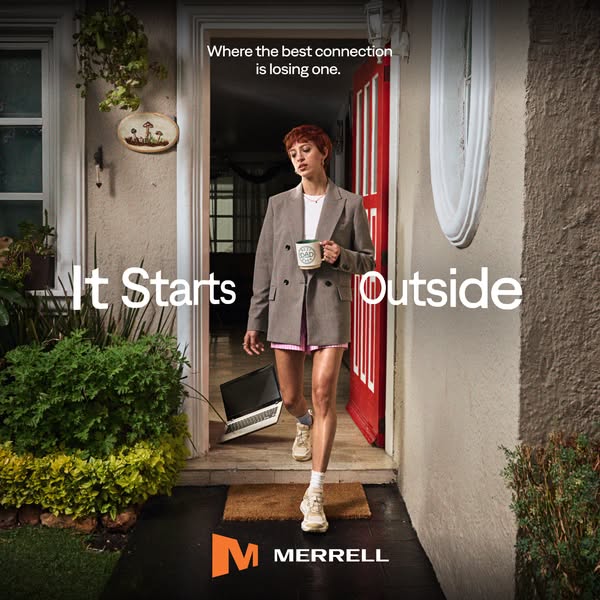 Woman in grey blazer walks out of a front door with a mug, laptop left behind, wearing beige Merrell sneakers