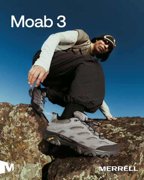 Crouching hiker puts a grey Merrell Moab 3 hiking shoe forward on a lichen-covered rock under a blue sky