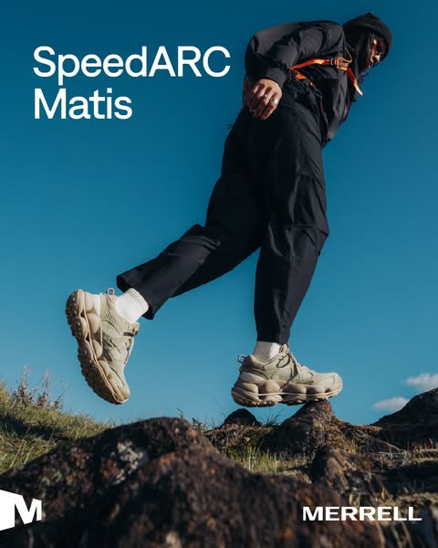 Hiker in black jumps across a rocky ridge in beige Merrell SpeedARC Matis trail shoes under a blue sky