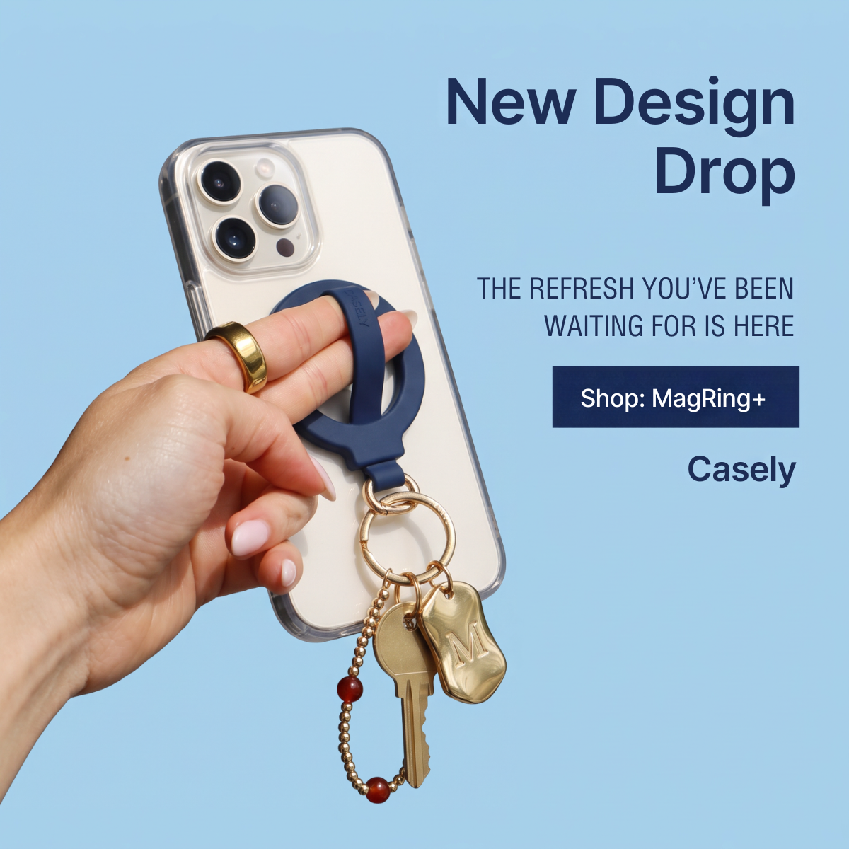 Hand holding iPhone with navy Casely MagRing+ grip and gold keys and charms on a light blue background